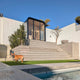 A modern outdoor architectural scene with a large concrete staircase leading up to a glass-walled pavilion, a swimming pool in the foreground, and a dog standing on green grass under a clear blue sky.