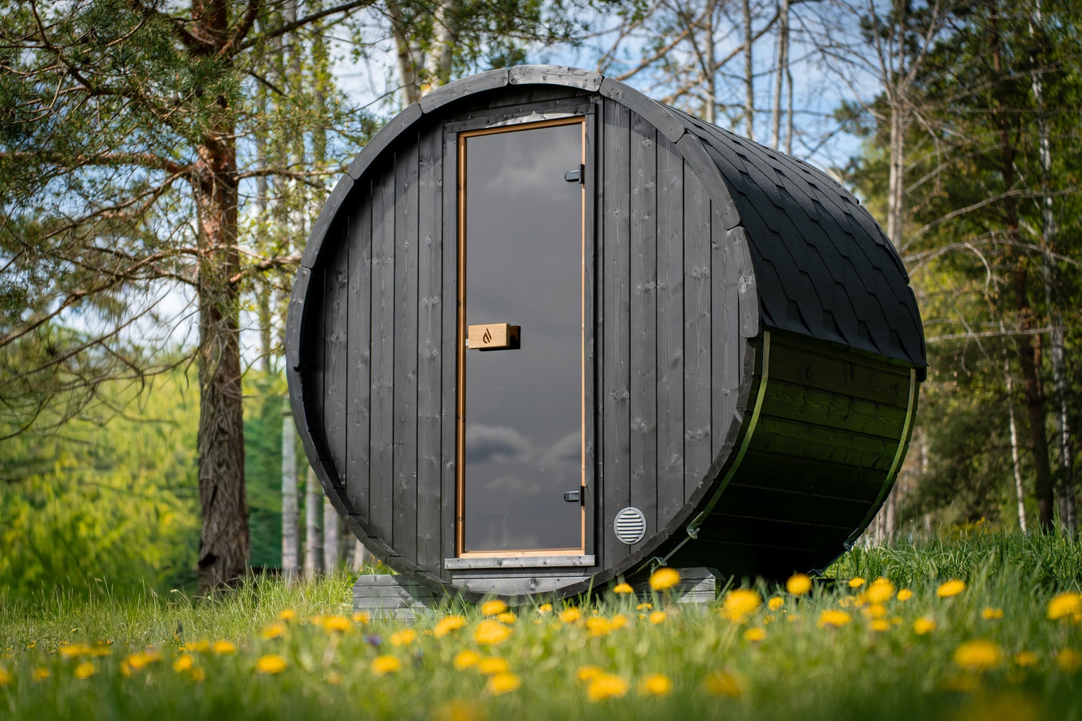 PlusLife Cove Barrel Sauna in black-stained Nordic spruce with a tall narrow tinted-glass door, in a garden