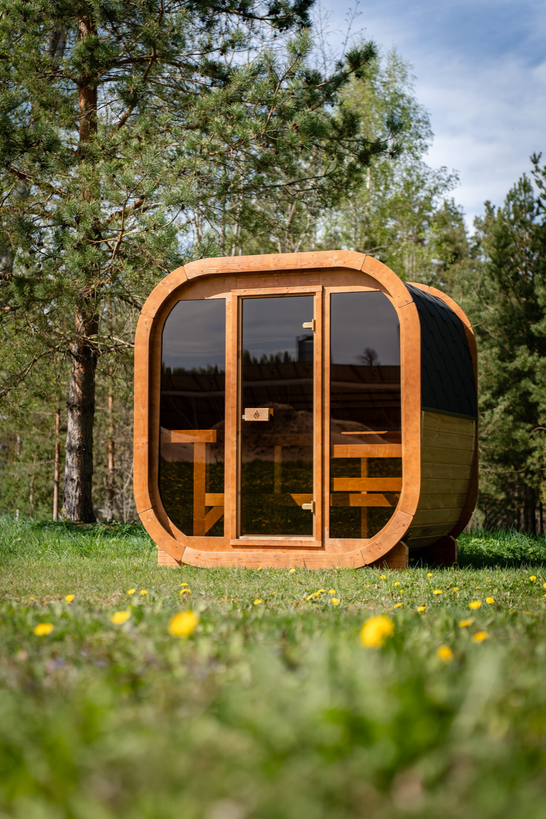PlusLife Max Traditional Sauna 2.0 seen from the front corner: rounded timber cabin with a glass front, a step up to the door and timber benches inside